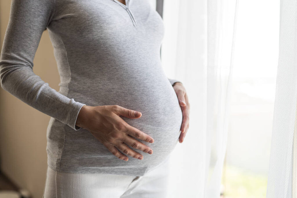 腹を手にしたグレーのスウェットシャツを着た妊婦のクローズアップは妊娠の最後の数ヶ月で窓の近くに立っています - 写真・画像