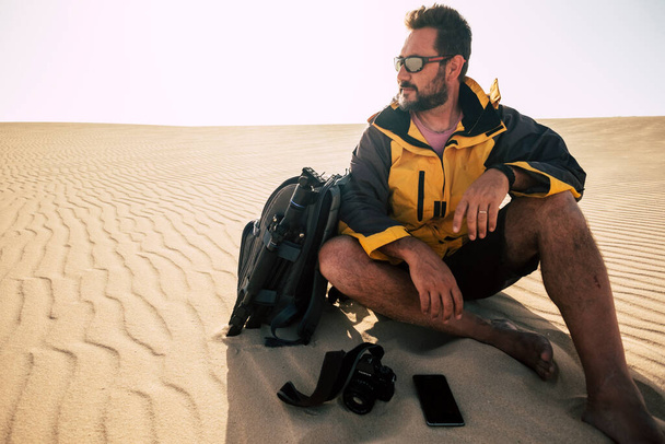 Νεαρός ενήλικας εξερευνά μόνος του αμμόλοφους της ερήμου με σακίδιο και φωτογραφική μηχανή. Φωτογραφία τοπίο δραστηριότητα εργασίας με γενειοφόρους άνδρες που κάθονται και ξεκουράζονται μετά το περπάτημα ξυπόλητοι σε αμμόλοφους - Φωτογραφία, εικόνα