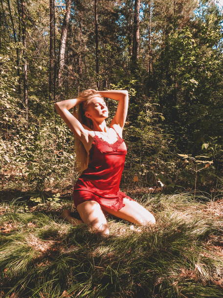 Sexy blonde woman with long hair in a red silk negligee posing in a summer coniferous forest, the concept of freedom and unity with nature, beauty and aesthetics of the female body, active lifestyle - 写真・画像
