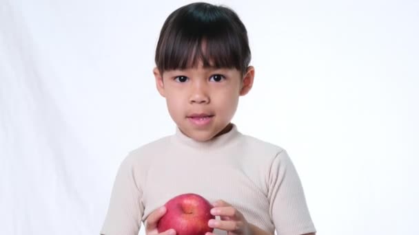 Szczęśliwa dziewczynka z jabłkiem. Cute Asian little girl jedzenia organiczne jabłko na białym tle w studio. Zdrowe odżywianie małych dzieci. - Materiał filmowy, wideo
