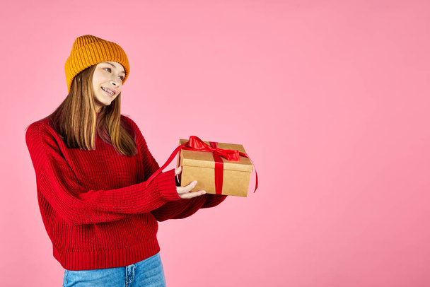 Młoda uśmiechnięta szczęśliwa hipsterka z bekonem dająca pudełka z okazji Nowego Roku, w czerwonym swetrze z dzianiny i żółtym kapeluszu. Prezenty świąteczne i trend mody zimowej. - Zdjęcie, obraz