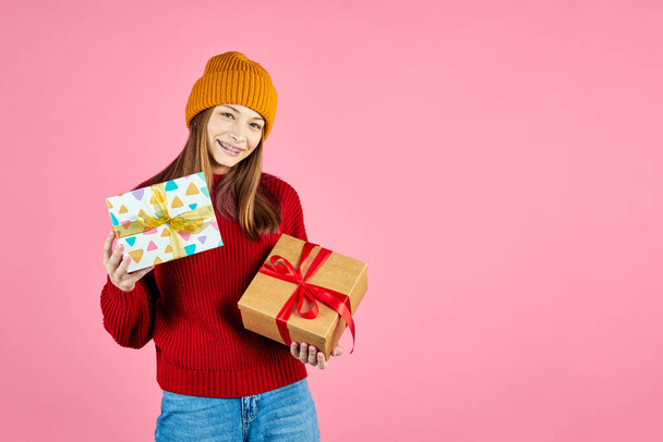 Young smiling happy hipster girl with baces holding present boxes celebrating new year, wearing red knitted sweater and yellow hat. Christmas gifts and winter fashion trend. - Fotoğraf, Görsel