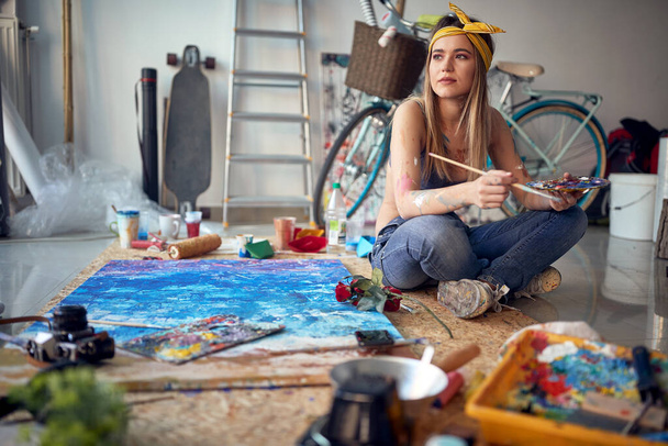 A sexy and attractive female artist is thoughtfully sitting on the floor in a relaxed atmosphere in her studio and waiting for the inspiration while working on the painting. Art, painting, studio - Φωτογραφία, εικόνα