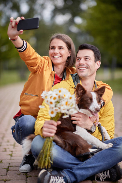 close up of young caucasian couple taking selfie with their dog in a lap, sitting on a path in park - Photo, Image