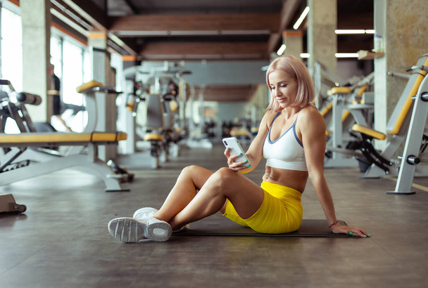 Mujer atlética hermosa joven usando teléfono inteligente mientras está sentado en la estera en el gimnasio - Foto, imagen