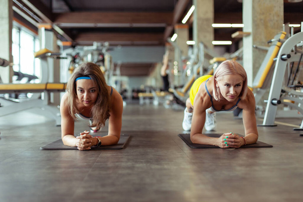 Две молодые атлетические женщины практикуют доски упражнения во время тренировки выносливость в тренажерном зале. Совместное обучение. Здоровый образ жизни - Фото, изображение