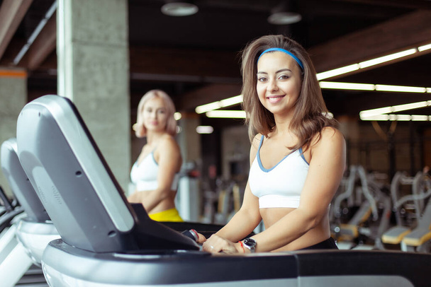 Gezond lachende jonge vrouw aan het trainen op een loopband in een moderne fitnessruimte. Gezondheid en fitness, cardiotraining - Foto, afbeelding