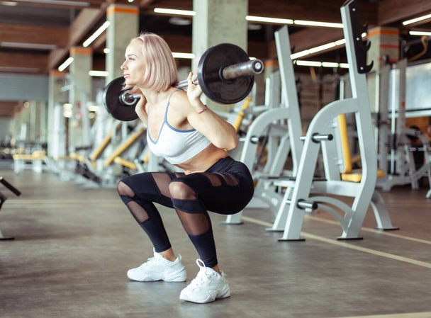 Athletic beautiful woman squats with barbell on her shoulders in the gym. Healthy lifestyle - Фото, зображення