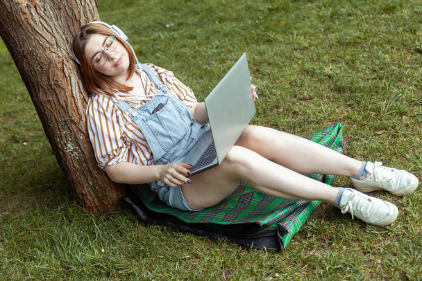 Jonge lachende vrouw met laptop en hoofdtelefoon in slaap leunend op een boom in het park - Foto, afbeelding