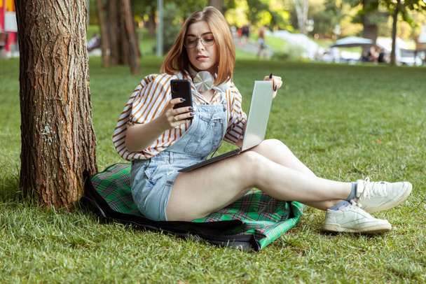 Joven mujer sonriente con portátil usando el teléfono apoyado en un árbol en el parque - Foto, imagen