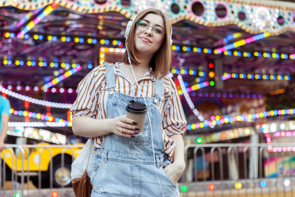 Mujer elegante lindo en gafas y overoles de mezclilla, escuchando música en los auriculares, sosteniendo la taza de café en un parque de atracciones - Foto, imagen