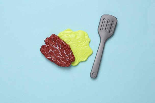 Plastikowe mięso, liść sałatki i widelec na niebieskim pastelowym tle. Widok góry. Minimalna koncepcja żywności - Zdjęcie, obraz