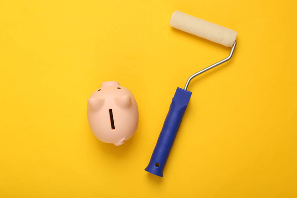 Paint roller with piggy bank on yellow background. Home renovation concept. Top view  - Фото, зображення
