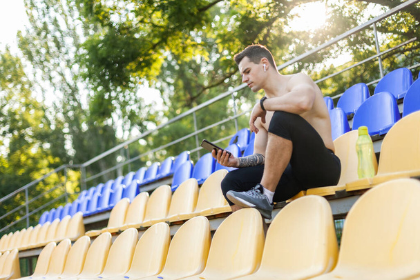Спортсмен відпочиває на стільці на трибуні стадіону і використовує телефон
 - Фото, зображення