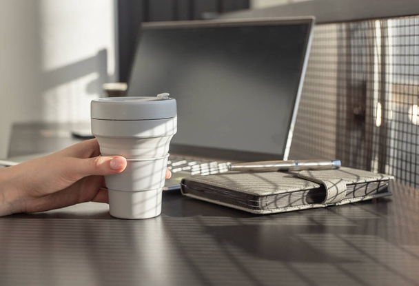 Женщина держит чашку на вынос и работает над ноутбуком. Бизнесмен или студентка, сидящая за столом с планировщиком и компьютером. Перерыв на кофе.  - Фото, изображение