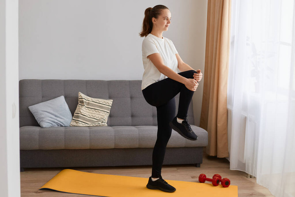 Side view of attractive woman wearing white t shirt and black leggins doing sport exercises at home, standing with one leg lift, thighs workout, warming up training muscles. - Fotó, kép