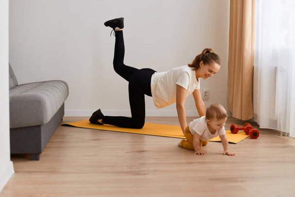 Image of positive adorable woman wearing white t shirt and black leggins doing sport exercises at home, doing one legged plank pose bow leg with her baby, training muscle flexibility. - Foto, Imagem
