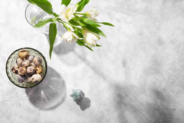 Quail eggs on the bowl and white spring flowers on the gray background, top view, free space for text. Festive easter card with copy space for text - Foto, imagen