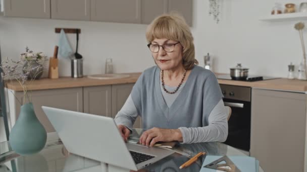 Retrato de una mujer caucásica mayor sentada en la mesa de la cocina con computadora portátil y copybooks y posando para la cámara mientras estudia en línea desde casa - Imágenes, Vídeo