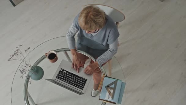 Top-Down-Aufnahme einer Seniorin, die am Laptop tippt, während sie an einem Glastisch mit Textbüchern, Kaffee und einer Vase mit Blumen sitzt. Arbeit von zu Hause aus und Fernstudium - Filmmaterial, Video