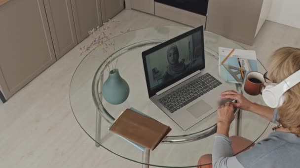 Ujęcie z góry na dół dojrzałej kobiety w słuchawkach bezprzewodowych siedzącej przy stole w kuchni i słuchającej nauczycielki w hidżabie podczas lekcji online przez telefon wideo na laptopie - Materiał filmowy, wideo