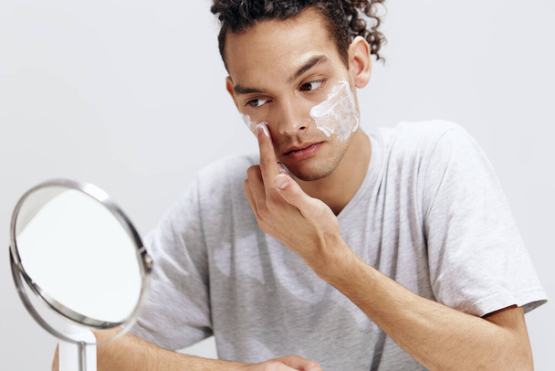 chlap s kudrnaté vlasy nanášení krém na obličej a při pohledu do zrcadla kosmetologie světlo pozadí - Fotografie, Obrázek