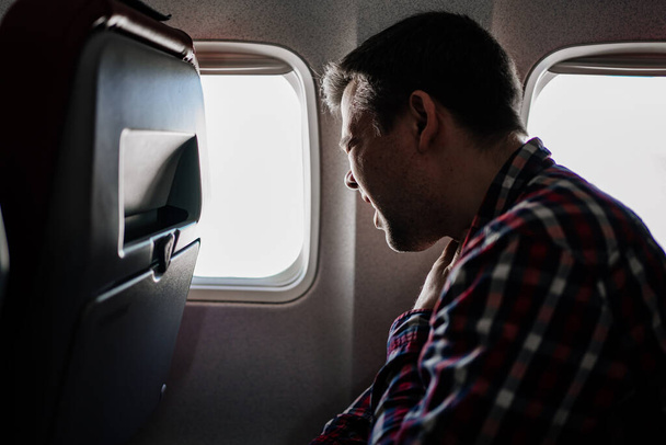 a man in a plaid shirt sneezes on the plane in a window seat.  - Фото, изображение