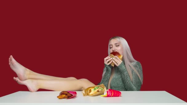 Sevimli bir kız bacakları masada bir sandalyeye oturur ve kırmızı arka planda hamburger yer. Diyet olsun. Sağlıklı ve sağlıksız gıda kavramı. fast food - Video, Çekim