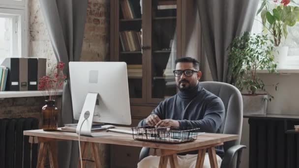 Alegre hombre de Oriente Medio usando la computadora en el escritorio, luego posando para la cámara y sonriendo mientras trabaja en la oficina en casa - Metraje, vídeo
