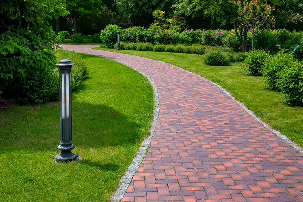 iron ground lantern garden lighting of park curved path paved with stone tiles in park among plants, evergreen bushes and foliage trees surrounded by eco friendly green lawn on sunny day, nobody. - Photo, image