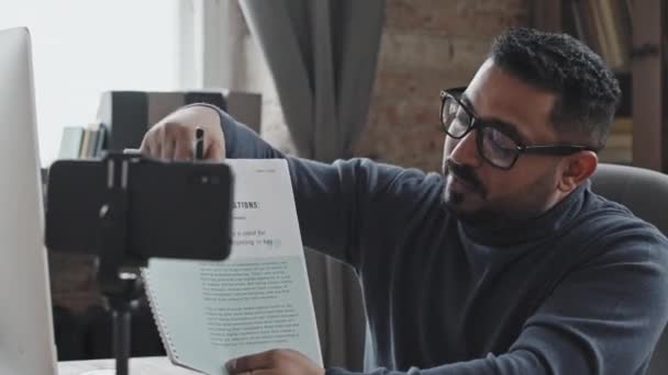 Tutor masculino de Oriente Medio explicando la tarea en el libro de trabajo en la cámara del teléfono inteligente en poder del trípode mientras da una lección en línea a través de videollamada - Metraje, vídeo