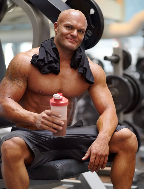 Genießen Sie einen Proteinshake nach dem Training. Porträt eines männlichen Bodybuilders, der einen Proteinshake hält, während er auf einer Hantelbank im Fitnessstudio sitzt. - Foto, Bild