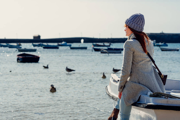 海を眺めながら漁船にもたれている毛むくじゃらの帽子を被った女性 - 写真・画像