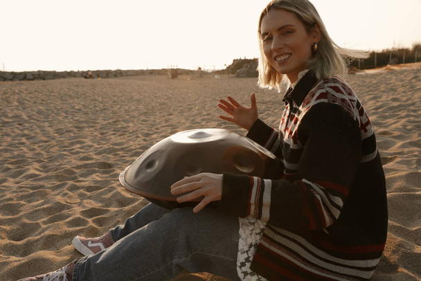 Woman plays relaxing music with hang drum handpan while sitting on the beach. - Фото, изображение
