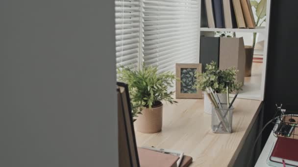 Arc shot van kamerplanten, digitale tablet, boeken, ingelijste foto, notitieblok en potloden in mesh houder op houten vensterbank in kantoor - Video
