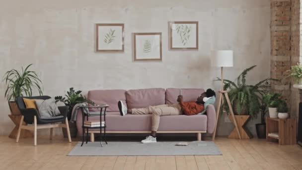 Amplia toma de chica afroamericana escuchando música con auriculares inalámbricos mientras está acostada en el sofá en casa y usando una tableta digital - Metraje, vídeo