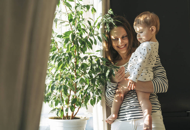 Счастливая мама молодая женщина и маленькая дочка смотрят на большое растение в комнате дома на жестком свете - Фото, изображение