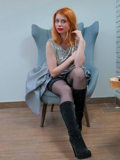 A red-haired attractive young woman in a blue evening dress with slender legs in black stockings, high-heeled boots, sitting in an armchair against the wall, fashion and style, evening dresses, meetings and romantic dates - Foto, immagini