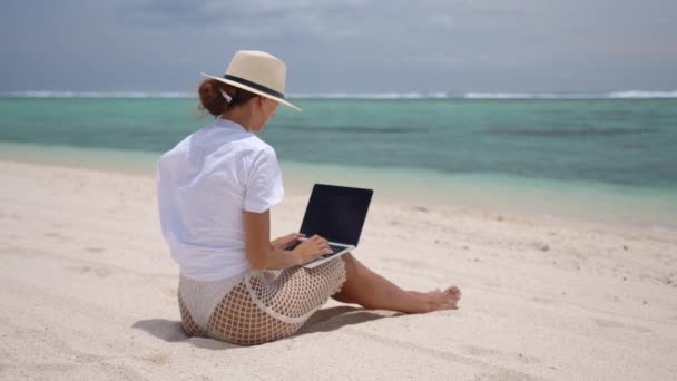 Sarı şapkalı başarılı bir iş kadını deniz kenarında dizüstü bilgisayarla çalışıyor. Yazın dışarıda bilgisayarda yazı yazan güzel bir bayan. Serbest çalışma, seyahat ve tatil kavramı. - Video, Çekim