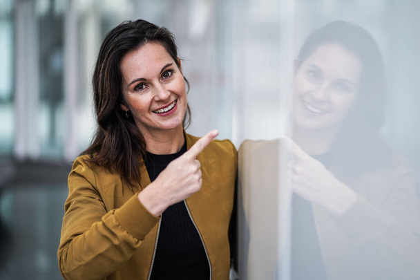 happy smiling casual business woman pointing finger at camera to address you directly for motivation surrounded by modern white indoor walls - Foto, immagini