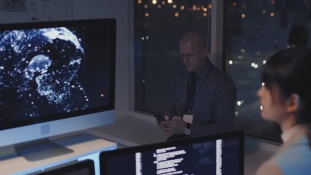 Medium of bold Caucasian man wearing eyeglasses, using smartphone, standing by window in corporate office at night, cropped Asian woman working in foreground - Footage, Video