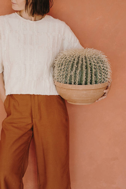 Junge schöne Frau in Ingwerhose und weißem T-Shirt hält Kaktus gegen Ingwerwand. Ästhetisch minimalistisches Modekonzept - Foto, Bild