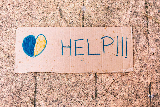 Signo de cartón que dice: '¡Ayuda!' junto a un corazón azul y amarillo, sobre un fondo urbano. Concepto de guerra de Ucrania, Rusia, avión, no a la guerra y detener la guerra. - Foto, Imagen
