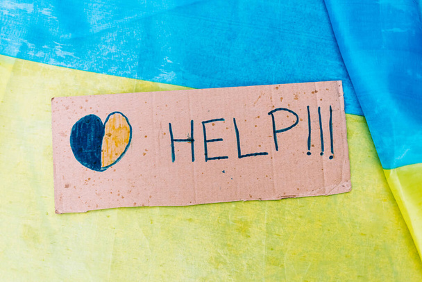 Πινακίδα από χαρτόνι που λέει: "Βοήθεια" δίπλα σε μια μπλε και κίτρινη καρδιά, στο φόντο των χρωμάτων της ουκρανικής σημαίας. Έννοια του πολέμου της Ουκρανίας, της Ρωσίας, inplane, όχι στον πόλεμο και να σταματήσει τον πόλεμο. - Φωτογραφία, εικόνα