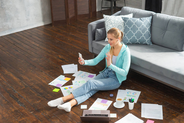 Young woman works with documents using smartphone and laptop while sitting on the floor at home. Student, entrepreneur or freelancer girl working or studying remotely via internet. - Photo, image