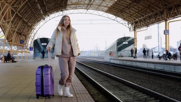 Een jonge vrouw in een warm pak en jas met een koffer wacht op een trein op het perron van het station - Video