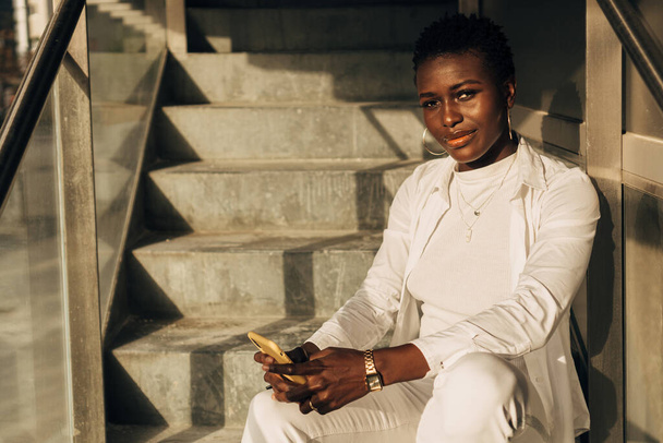 Stylish African American looking at the camera and holding a mobile phone while sitting on stairs. - Valokuva, kuva