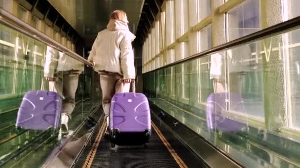 Молодая женщина в движущейся дорожке в аэропорту с фиолетовым чемоданом. - Кадры, видео