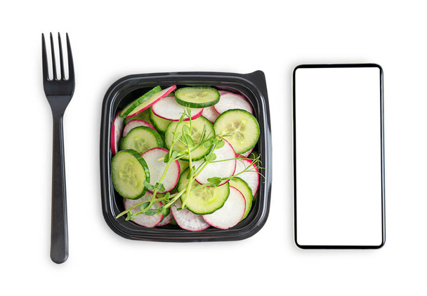 Vegetarian salad made of cucumber slices and radish served in black disposable plastic lunch box or container with fork and telephone with mock up isolated on white background. Food delivery concept - Foto, Imagen