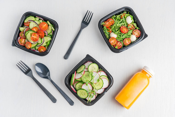 Variety of vegan vegetable salads made of cucumber slices, arugula and tomatoes served in black disposable lunch containers or boxes with orange juice bottle and tableware on white wooden background - Foto, Imagem